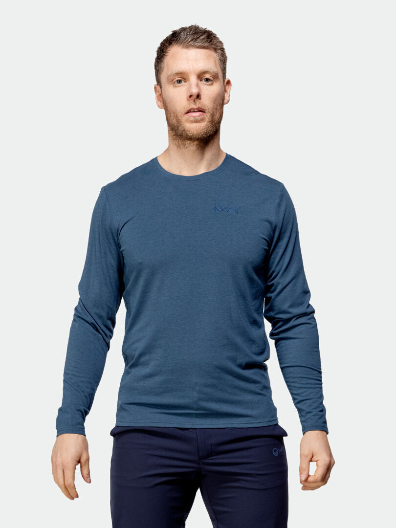 Mies seisoo kasvot menosuuntaan vaaleaa taustaa vasten, yllään pitkähihainen sininen paita ja tummat housut. Hänen kätensä ovat rentoina sivuillaan, ja hänen ilmeensä on neutraali.