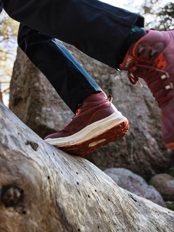 Henkilö, jolla on ruskeankeltainen hupparitakki, valkoinen neuletakki ja mustat käsineet, kävelee pilvisenä päivänä kaupungin kadun poikki vaaleiden rakennusten lähellä.