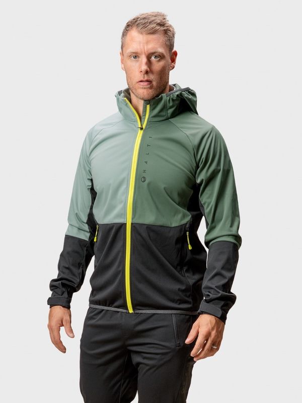 Mies seisoo kasvot menosuuntaan pukeutuneena vihreämustaan hupulliseen takkiin, jossa on kirkkaan keltainen vetoketju, ja mustiin housuihin. Takissa on korkea kaulus ja näkyvät saumojen yksityiskohdat. Tausta on vaaleanharmaa.