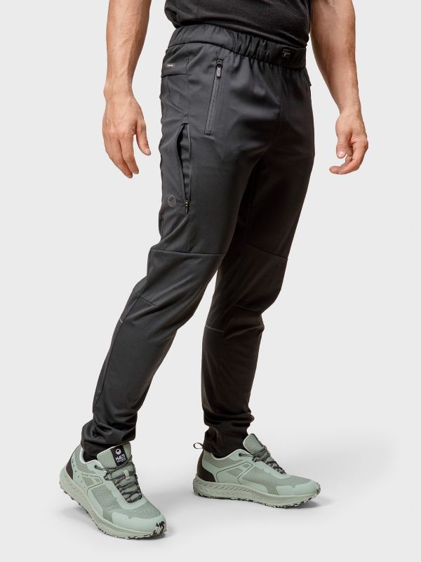 Henkilö, jolla on mustat urheiluhousut, joissa on vetoketjulliset taskut, ja vaaleanvihreät lenkkarit, seisoo vaaleanharmaalla taustalla.