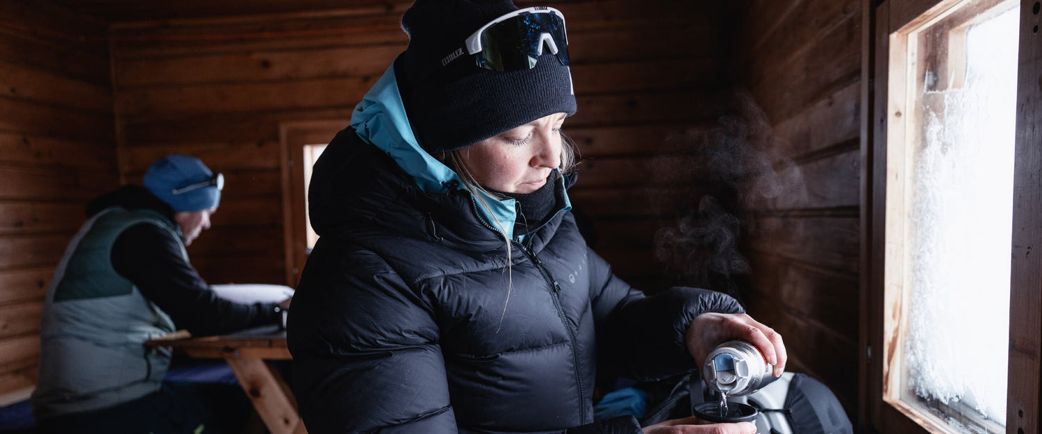 Mustaan talvitakkiin ja pipoon pukeutunut henkilö kaataa kuumaa juomaa termospullosta puisessa mökissä, taustalla pöydän ääressä istuu toinen henkilö.