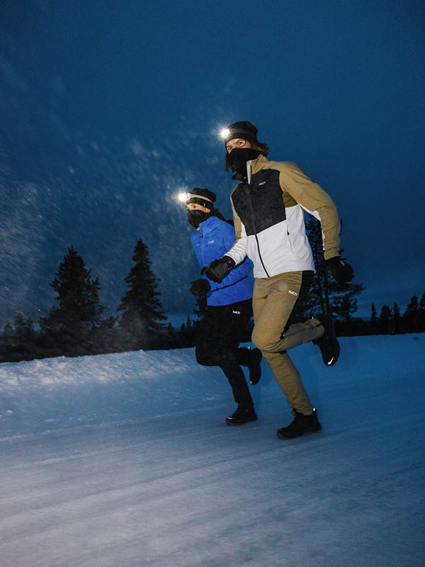 Kaksi otsalamppuihin, talvitakkeihin ja kasvosuojiin pukeutunutta ihmistä juoksee yöllä lumisella polulla, jonka taustalla on puita ja tumma taivas.