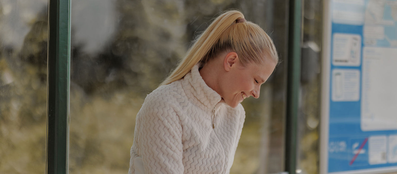 Nainen, jolla on pitkät vaaleat hiukset poninhännässä ja jolla on yllään valkoinen kuvioitu pusero, istuu ja katsoo alaspäin hymyillen. Hän on bussipysäkillä, jonka taustalla näkyy julisteita.