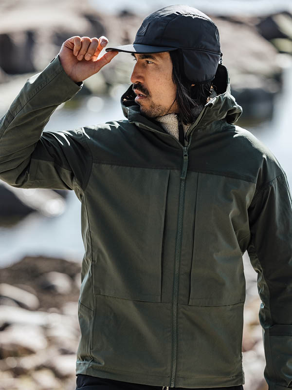 Tummanvihreään takkiin ja mustaan lippalakkiin pukeutunut henkilö seisoo ulkona ja pitää lippalakin lippalakkia. Taustalla on epäselviä kiviä ja vettä.