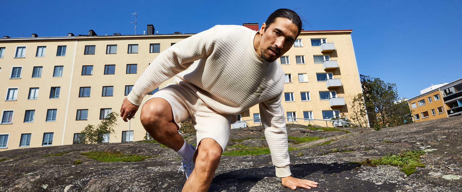 Kermanväriseen villapaitaan ja shortseihin pukeutunut mies kyyristelee ulkona kivisellä pinnalla, taustalla beige kerrostalo ja kirkkaansininen taivas.