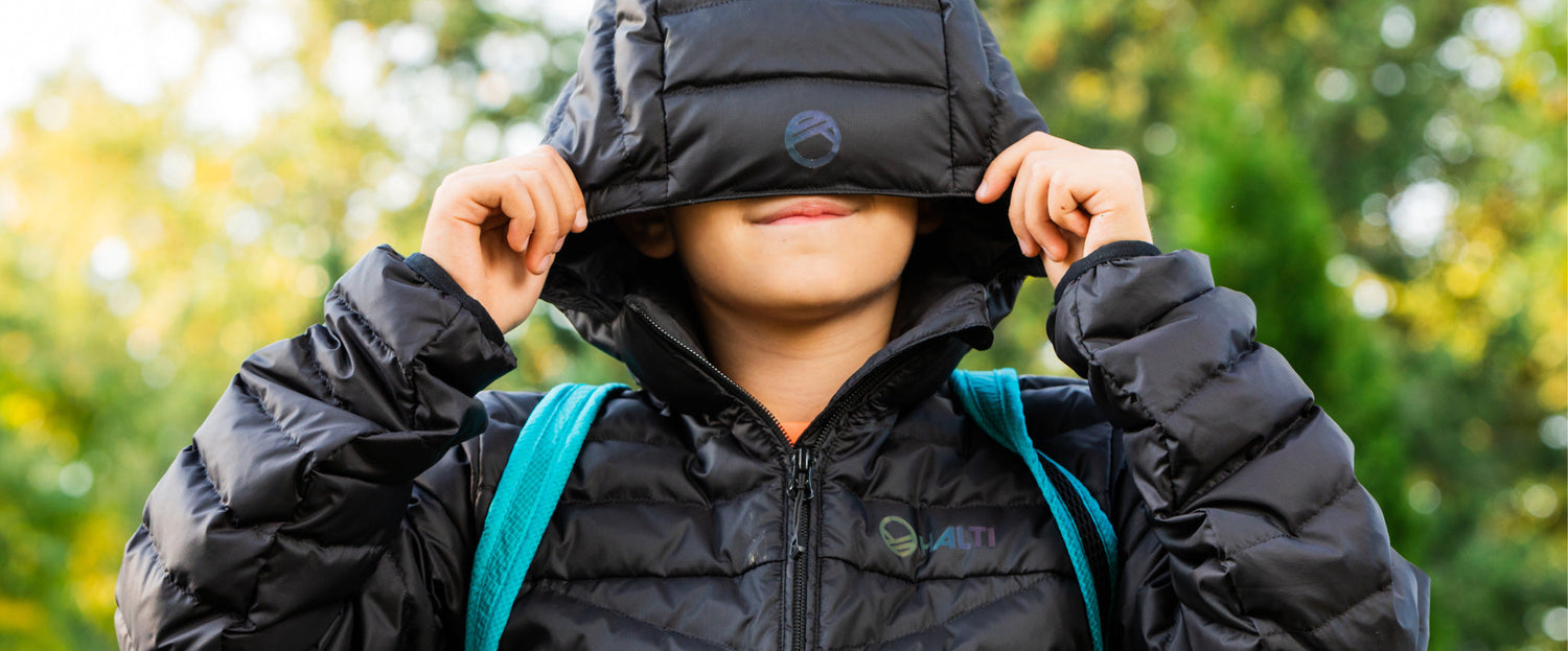 Henkilö, jolla on yllään musta puserotakki ja tiilenpunainen reppu, vetää hupun alas silmiensä päälle ja peittää osittain kasvonsa. Vihreät puut näkyvät epäselvässä taustassa.