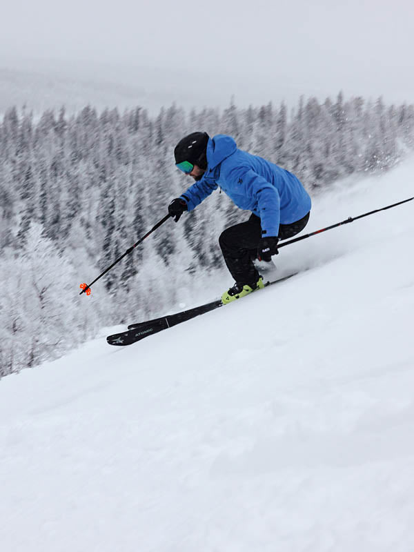 Siniseen takkiin, mustiin housuihin ja kypärään pukeutunut henkilö hiihtää alaspäin lumisella rinteellä suksisauvat kädessään, taustalla näkyy lumen peittämiä puita.