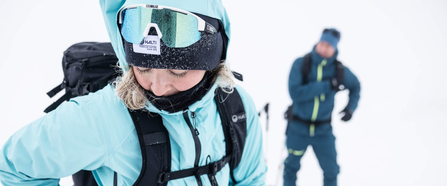 Siniseen talvitakkiin, lumilaseihin ja reppuun pukeutunut henkilö seisoo lumessa, ja taustalla on toinen henkilö, jolla on samanlaiset talvivarusteet.