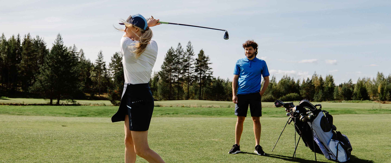 Valkoiseen paitaan ja mustaan hameeseen pukeutunut nainen heiluttaa golfmailaa nurmikentällä, kun siniseen paitaan pukeutunut mies seisoo lähellä golfpussin vieressä, taustalla puita.