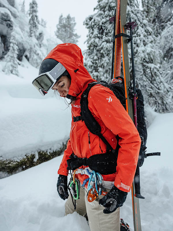 Henkilö, jolla on reppu, otsalamppu ja talvitakki, seisoo ulkona yöllä lumisessa maisemassa, jonka taustalla näkyy puita ja kukkuloita.