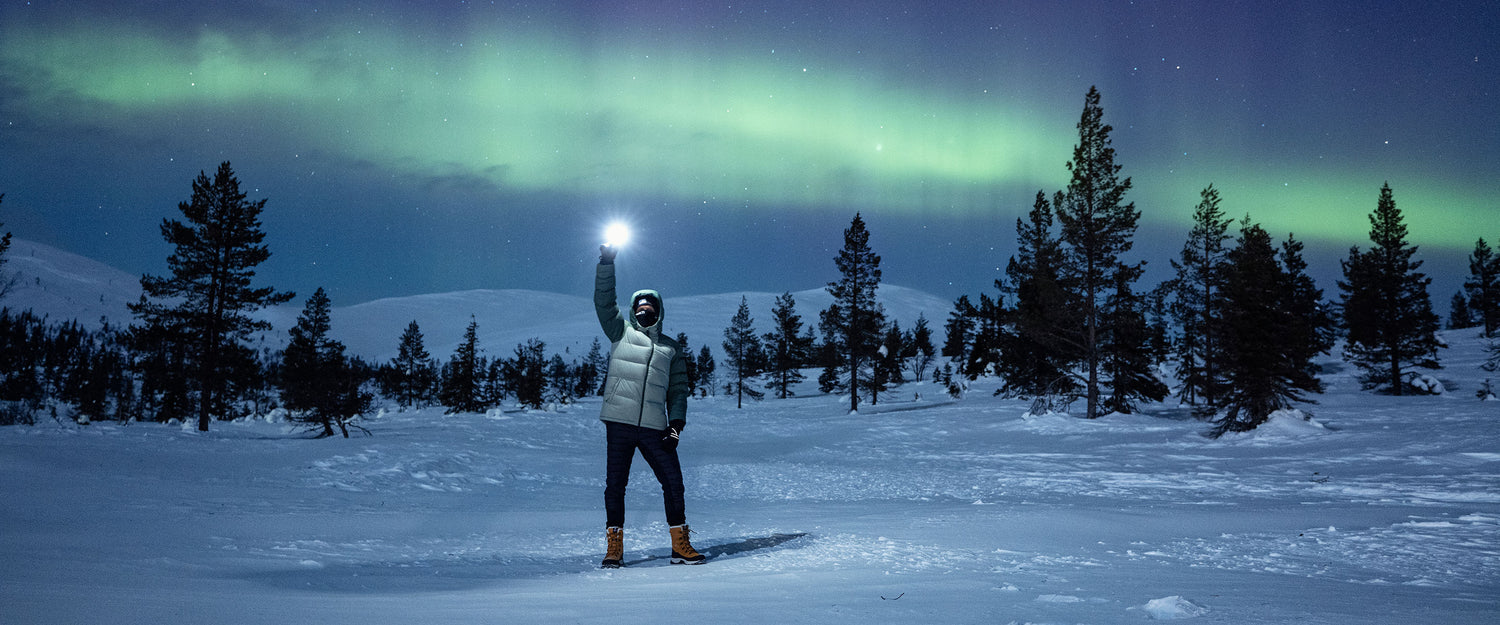 Talvivaatteisiin pukeutunut henkilö seisoo yöllä lumipeitteisellä maastolla ja pitelee kirkasta valoa, ja taustalla näkyvät vihreät revontulet ja männyt.