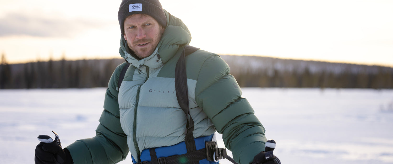 Henkilö, jolla on vihreä puskuritakki, siniset hiihtohousut, hanskat ja musta pipo, on ulkona lumisessa maisemassa, jonka taustalla on puita, ja hänellä on suksisauvat kädessään.
