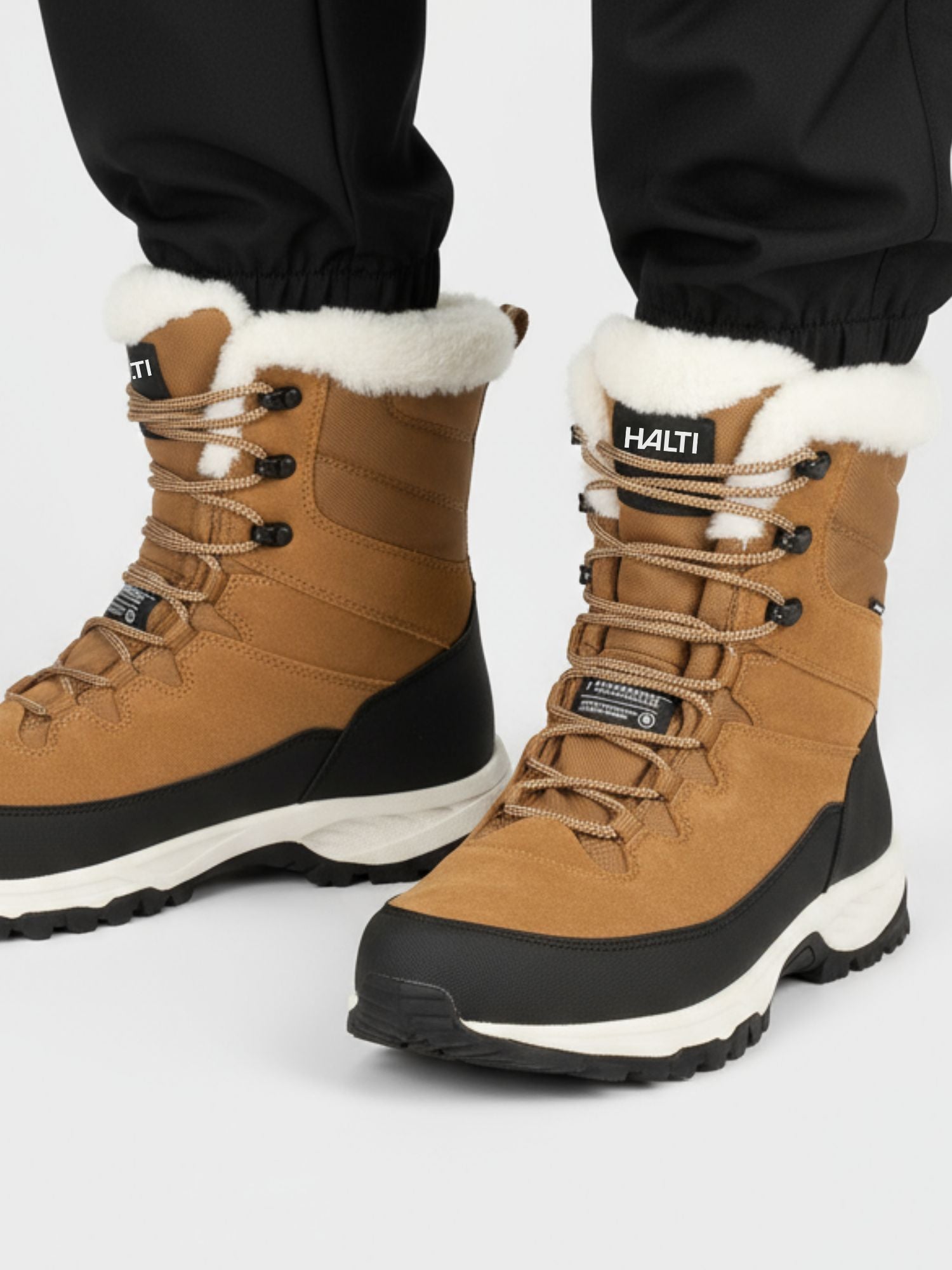 Henkilöllä on ruskeanruskeat nauhalliset talvikengät, joissa on valkoinen fleecevuori ja paksut mustavalkoiset pohjat, sekä mustat housut. Saappaiden kielessä on HALTI-logo.