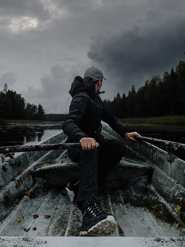 Tummiin vaatteisiin ja lippalakkiin pukeutunut henkilö istuu soutuveneessä rauhallisella vesialueella ja soutaa airoilla. Taivas on pilvinen ja sitä ympäröi tiheä puusto.