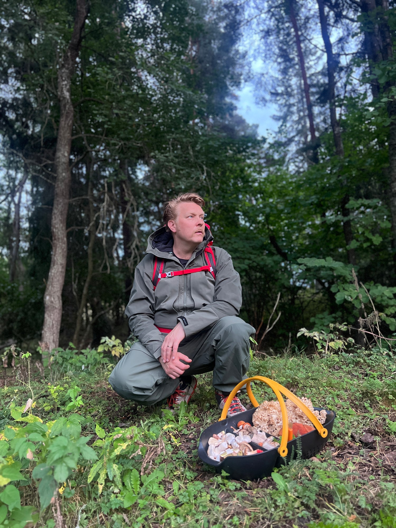 Ulkovaatteisiin pukeutunut henkilö polvistuu metsässä ruohikolla sieniä täynnä olevan korin vieressä.