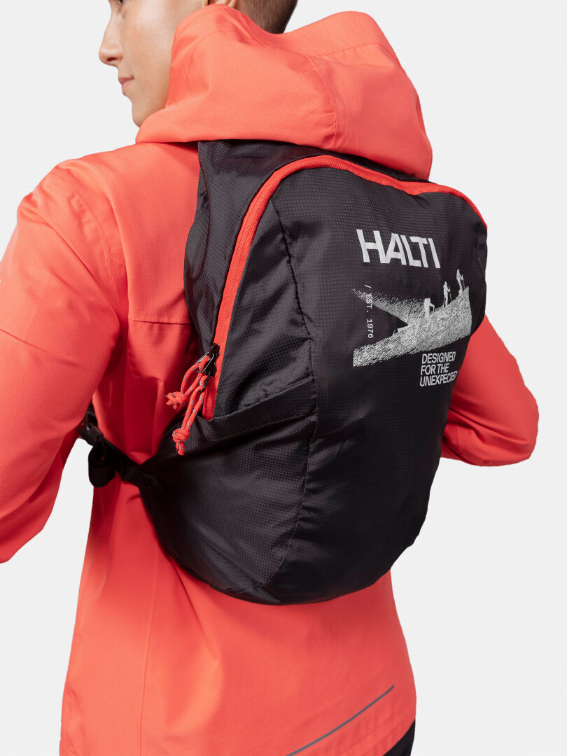 Henkilö korallinvärisessä takissa ja mustassa Halti Streetpack Reppu -kierrätysnylonista valmistetussa takissa vasemmalle päin.