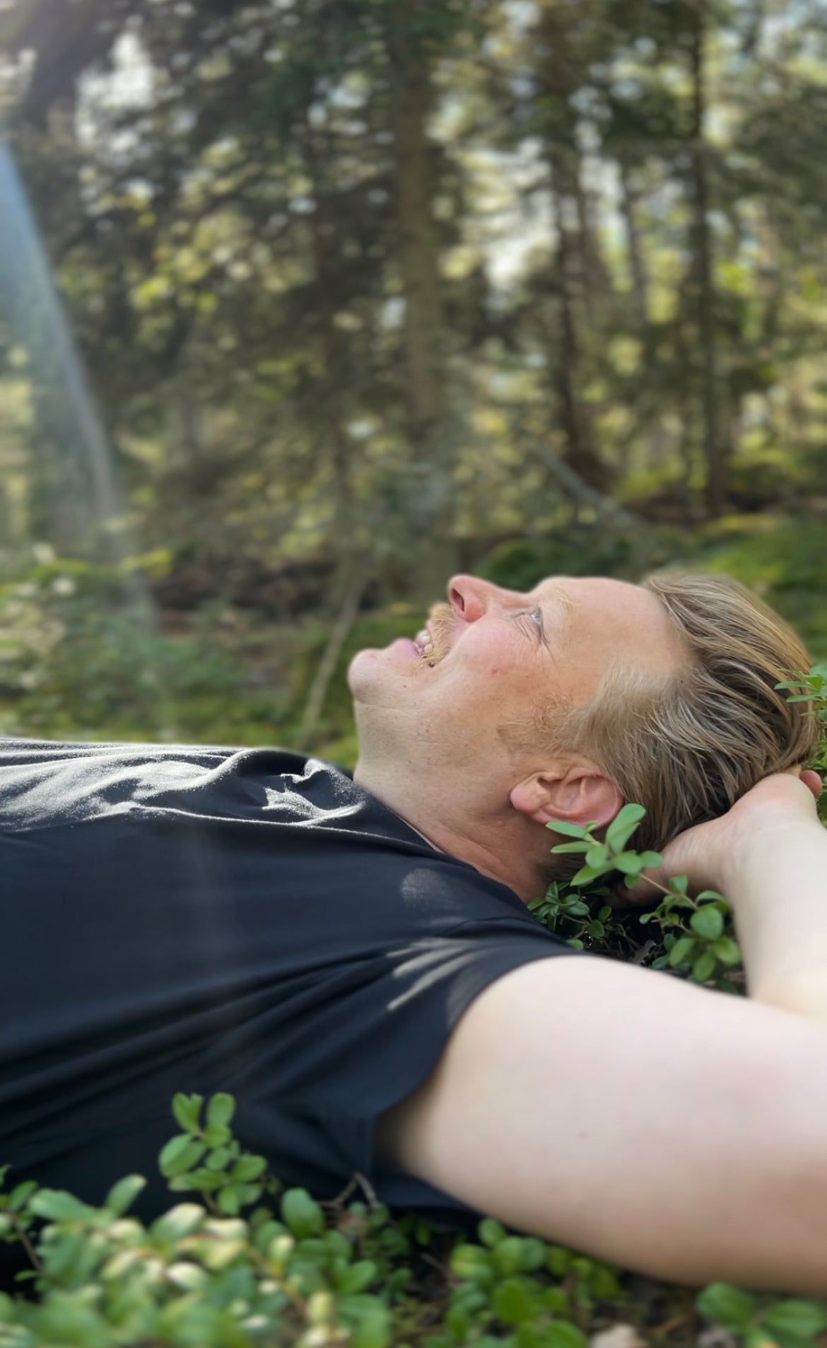 Henkilö makaa selällään metsässä, hymyilee ja katsoo ylöspäin, kun auringonvalo virtaa puiden läpi.