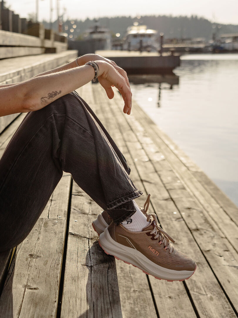 Tummissa farkuissa ja beigenvärisessä Halti Rapid Lenkkari Naisten -puvussa oleva henkilö istuu puisella laiturilla veden äärellä, veneet takana.