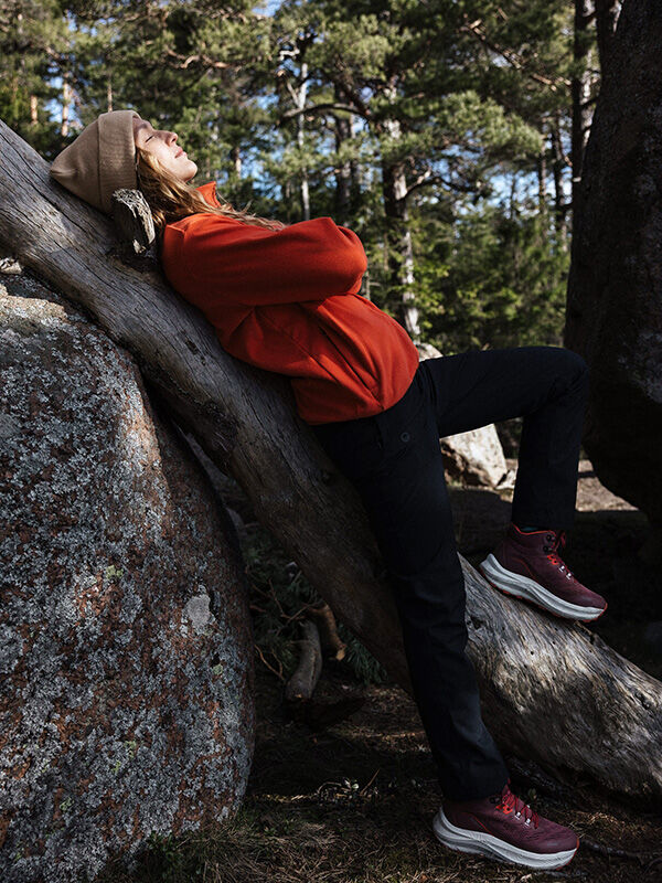 Henkilö, jolla on yllään punainen takki, vaaleanruskea hattu ja Halti Rapid Mid DX -naisten ulkoilukengät, nojaa metsässä kaatuneeseen puuhun silmät kiinni.