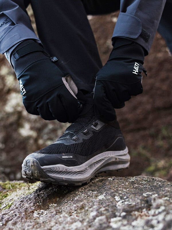 Henkilö, jolla on mustat hanskat, sitoo Halti Rapid Mid Freelock DX Ulkoilukenkä Unisexin nauhoja kalliolla ulkona.