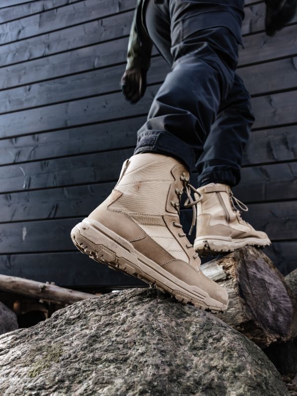 Tummiin housuihin pukeutunut henkilö astuu kiville, yllään Halti Regis DX Vaelluskenkä Miesten tumman puuseinän vieressä.