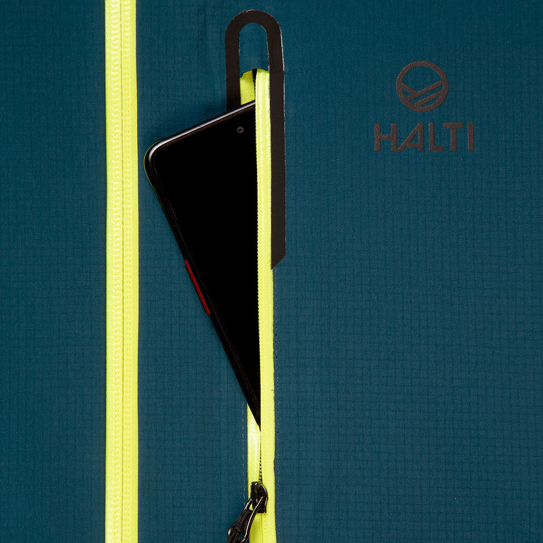 Sinisen Halti Alpine 3L DX Kuoritakki Unisexin vetoketjullisesta keltaisesta taskusta kurkistaa puhelin, merkin logo näkyvissä.