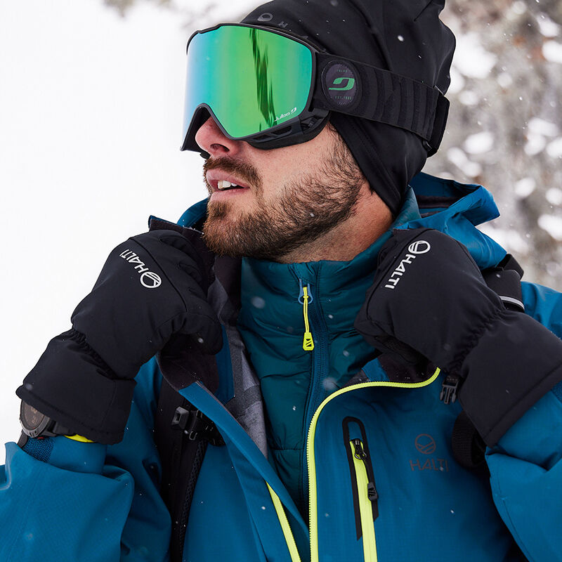 Hiihtolaseihin ja mustiin hanskoihin pukeutunut mies käyttää Halti Alpine 3L DX Kuoritakki Unisex sinistä takkia ulkona lumessa.