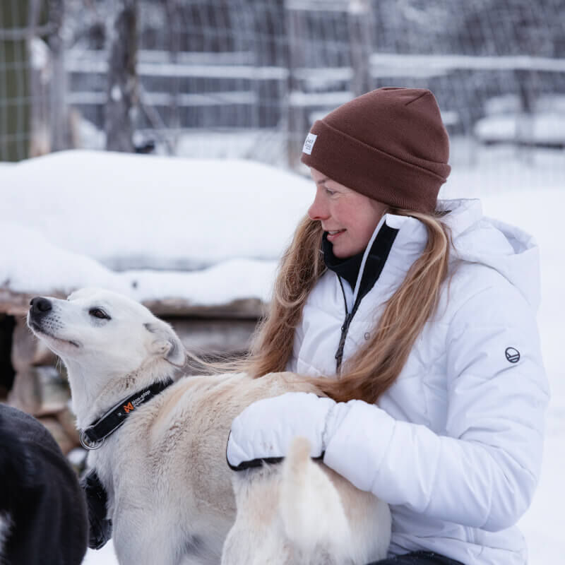 Henkilö Halti Mellow Puffer Laskettelutakki Naisten -koirassa silittää valkoista koiraa ulkona lumessa aidan vieressä.