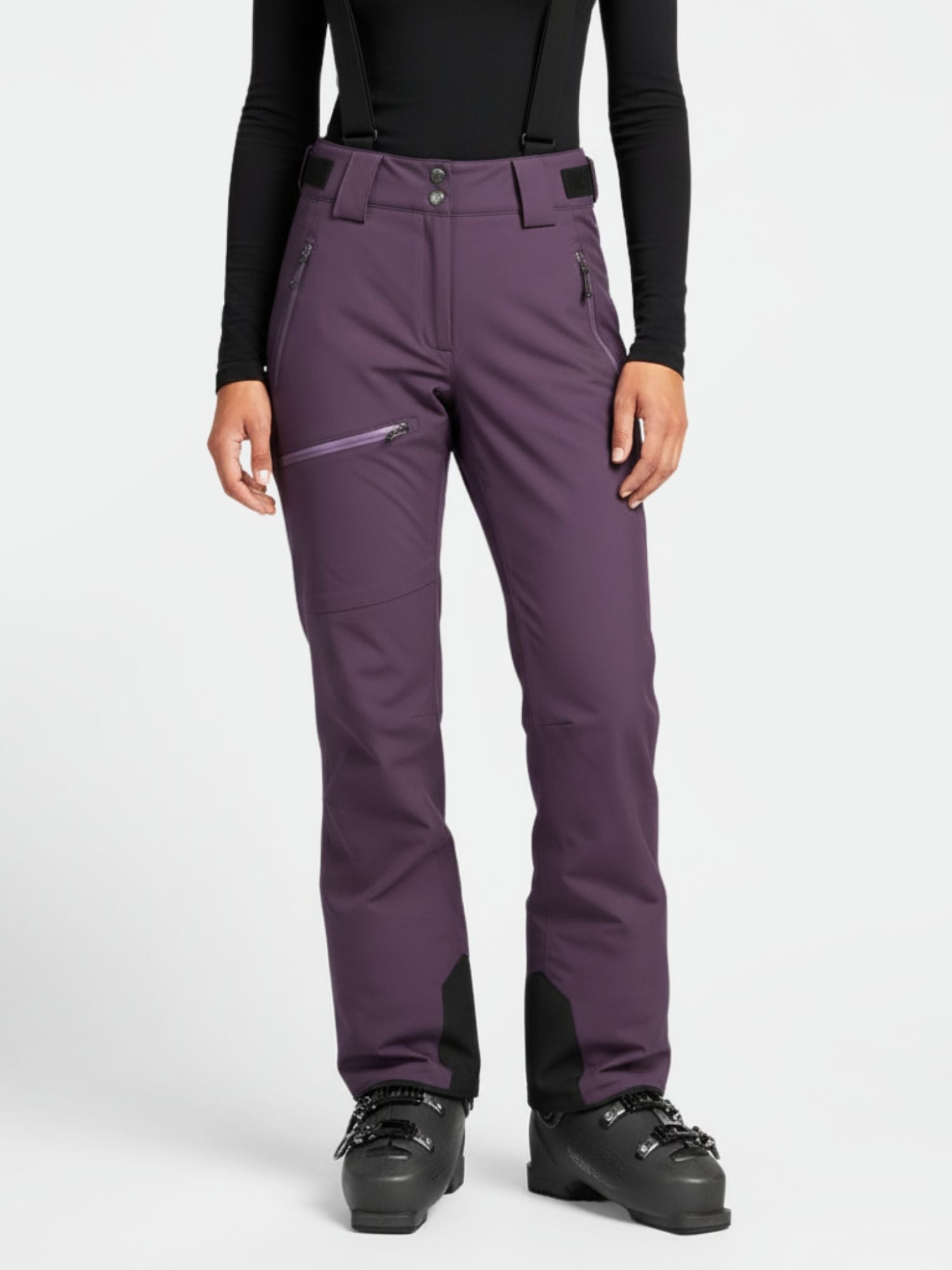 Henkilöllä on yllään violetit Halti Carvey DrymaxX Lasketteluhousut Naisten, mustat henkselit, musta toppi ja mustat hiihtokengät.