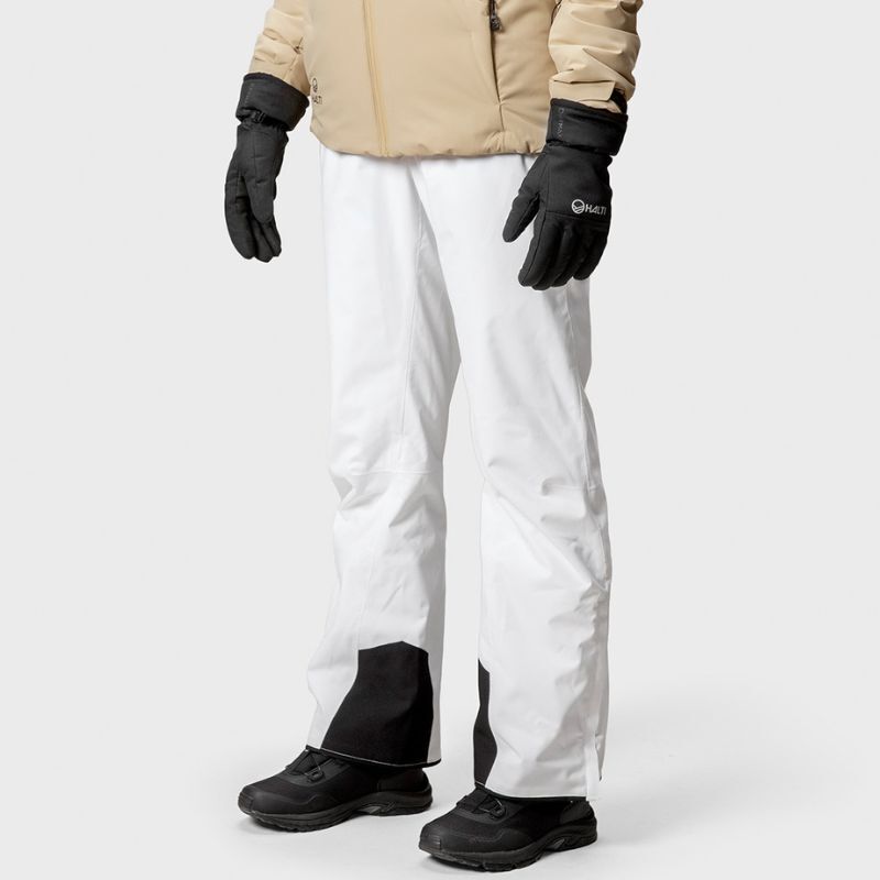 Henkilö valkoisissa Halti Trusty DrymaxX Lasketteluhousuissa, mustissa hanskoissa ja saappaissa, beigenvärisessä takissa yksiväristä taustaa vasten.