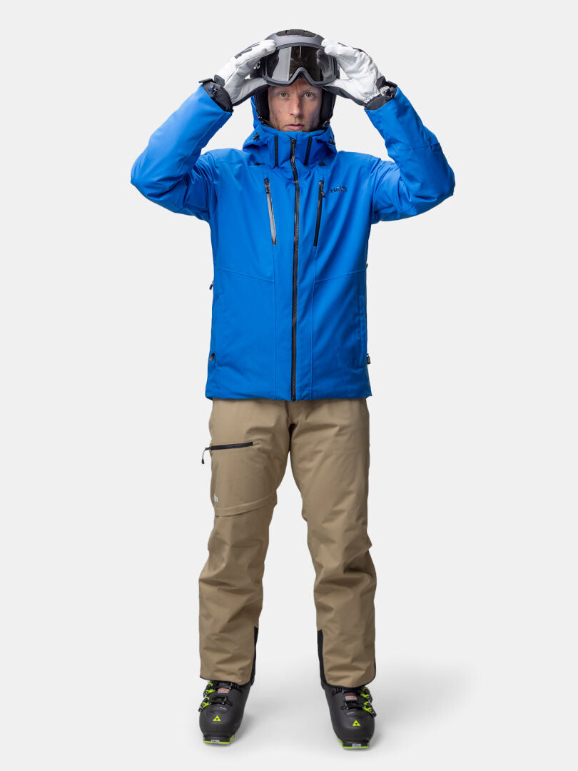 Henkilö, jolla on Halti Vertica DX Laskettelutakki Miesten, ruskehtavat housut ja hiihtokengät, säätää hiihtolaseja kasvot menosuuntaan.