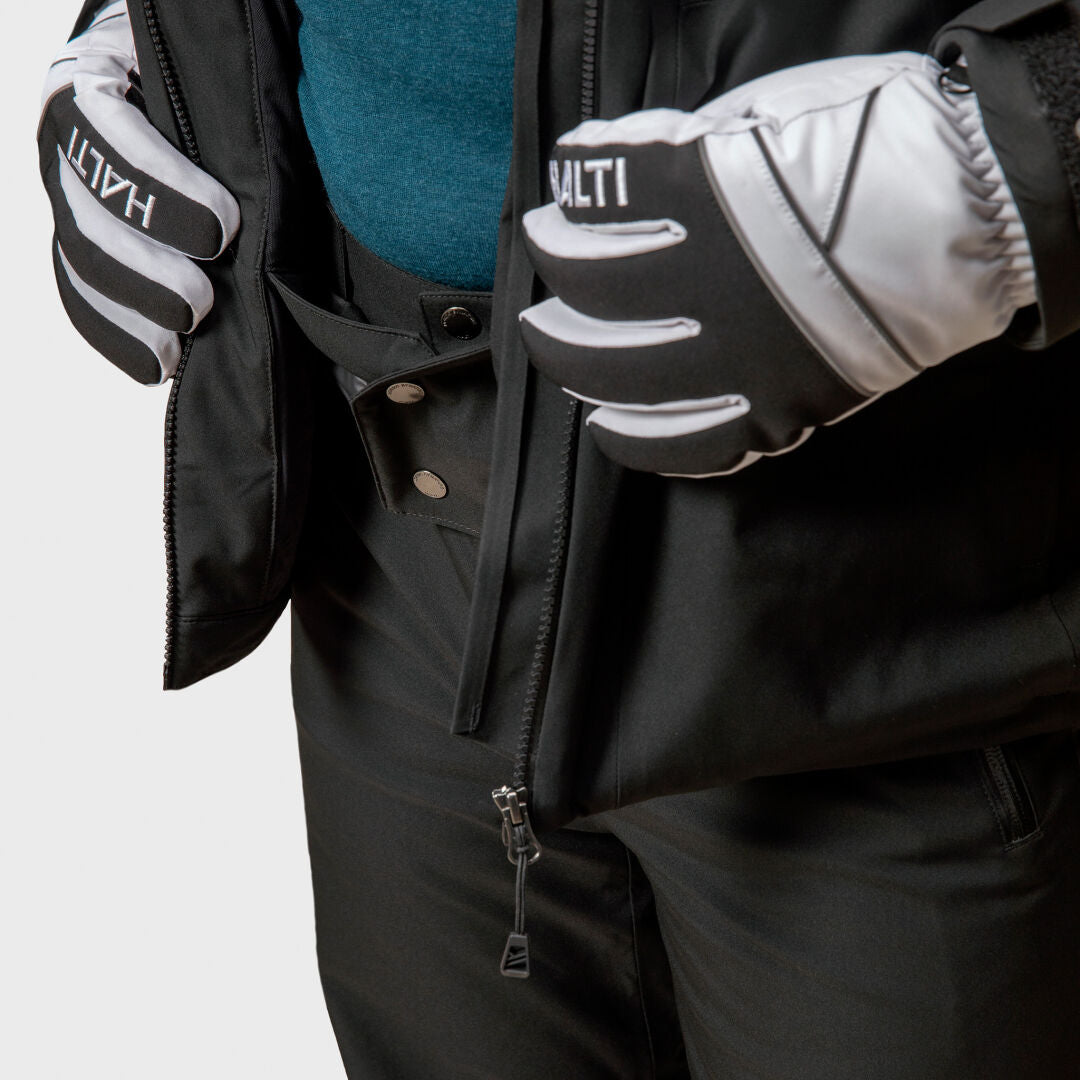 Henkilö vetoketjulla musta Halti Vertica DX laskettelutakki sinisen paidan päällä, yllään Halti-hanskat ja mustat lumihousut.