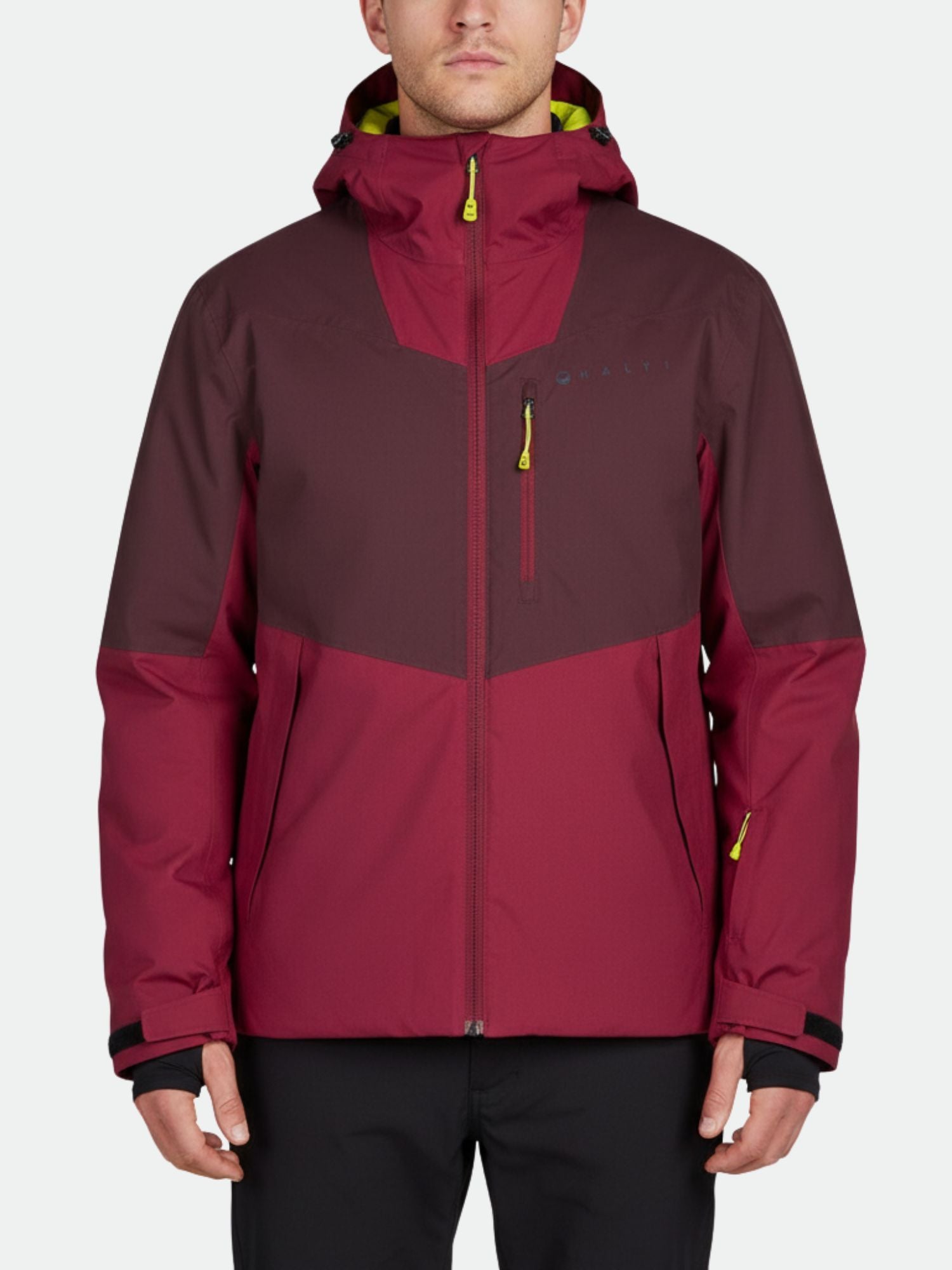 Henkilö, jolla on ruskeankeltainen Halti Wedeln DrymaxX Laskettelutakki Miesten, jossa on keltaisia korostuksia, seisoo tavallista taustaa vasten.