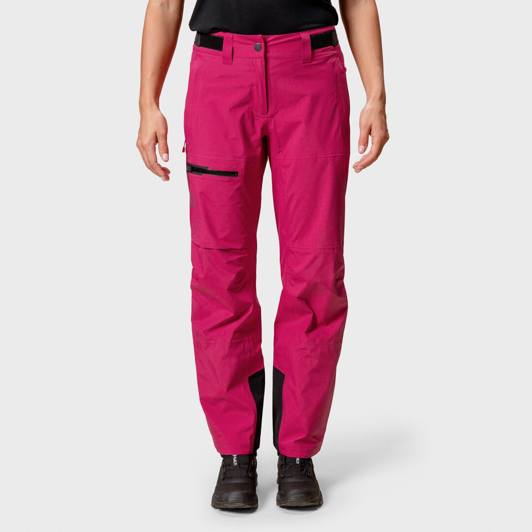 Henkilö, jolla on Halti Alpine 3L DX Kuorihousut Naisten, musta paita ja saappaat, seisoo vaaleaa taustaa vasten.