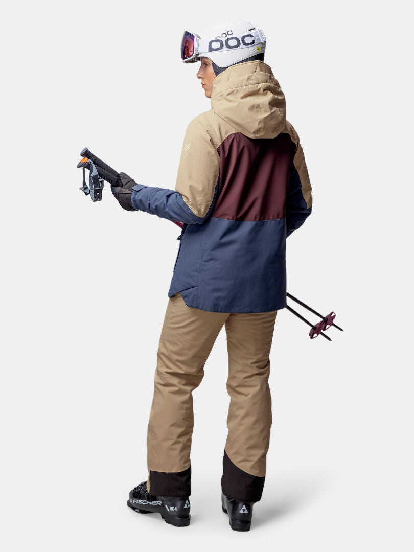 Ruskanruskeaan ja laivastonsiniseen Halti Gracer DX Laskettelutakki Naisten vaatteisiin pukeutunut henkilö seisoo hiihtovarusteiden kanssa, selkä kameraan päin.