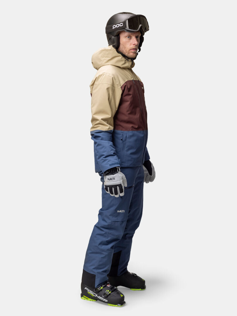 Mies seisoo sivuttain Halti Melvik DX Laskettelutakki Miesten sinisissä hiihtohousuissa, kypärässä, hanskoissa, saappaissa ja takissa.