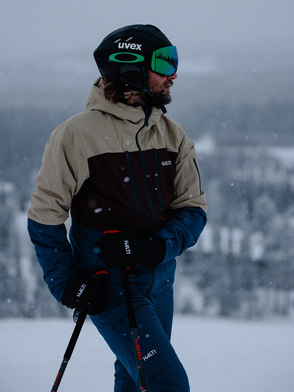 Halti Melvik DX Laskettelutakki Miesten hiihtäjä seisoo ulkona hiihtosauvojen kanssa, kun lunta sataa ympärillä.