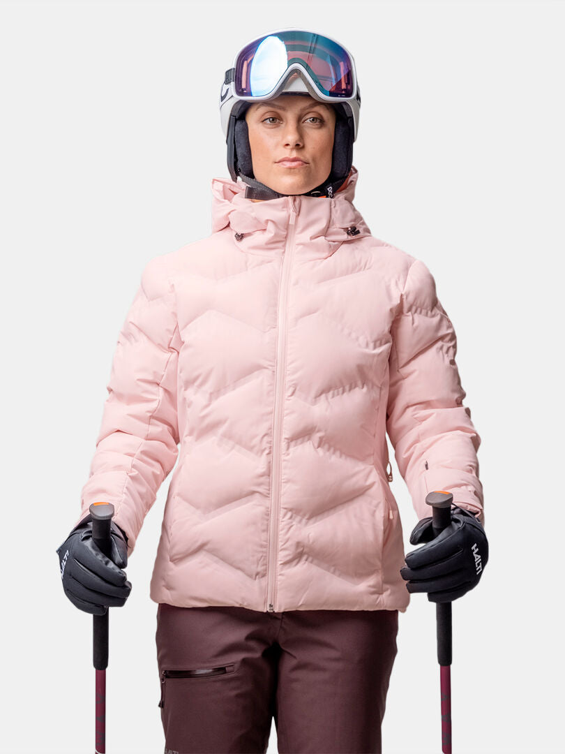 Vaaleanpunaisessa Halti Nordic Arcty II Laskettelutakki Naisten hiihtosuksilla hiihtävä henkilö pitelee hiihtosauvoja, päässään kypärä ja hiihtolasit.