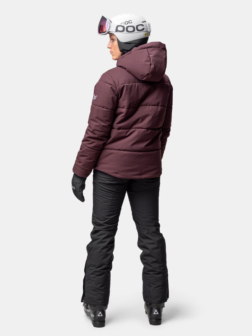 Henkilö, jolla on Halti Ascale Puffer Laskettelutakki, mustat housut, hanskat, valkoinen kypärä ja suojalasit, seisoo kasvot menosuuntaan.