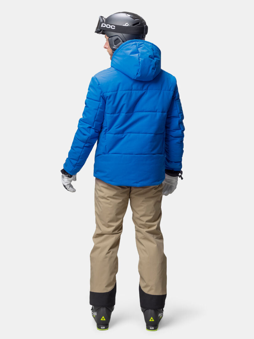 Henkilö sinisessä Halti Ascale Puffer Laskettelutakki Miesten ja beige housut kasvot poispäin valkoista taustaa vasten.