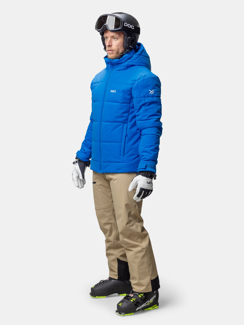 Henkilöllä on yllään sininen Halti Ascale Puffer Laskettelutakki Miesten, beige hiihtohousut, hiihtokengät, hanskat ja musta kypärä, jossa suojalasit.