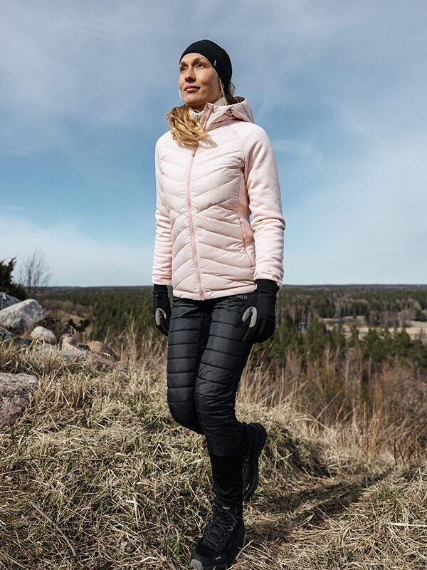 Vaaleanpunaiseen takkiin ja Halti Veidnes Kevyesti Topatut Housut Naisten vaatteisiin pukeutunut nainen kävelee nurmipolulla, jossa on puita.