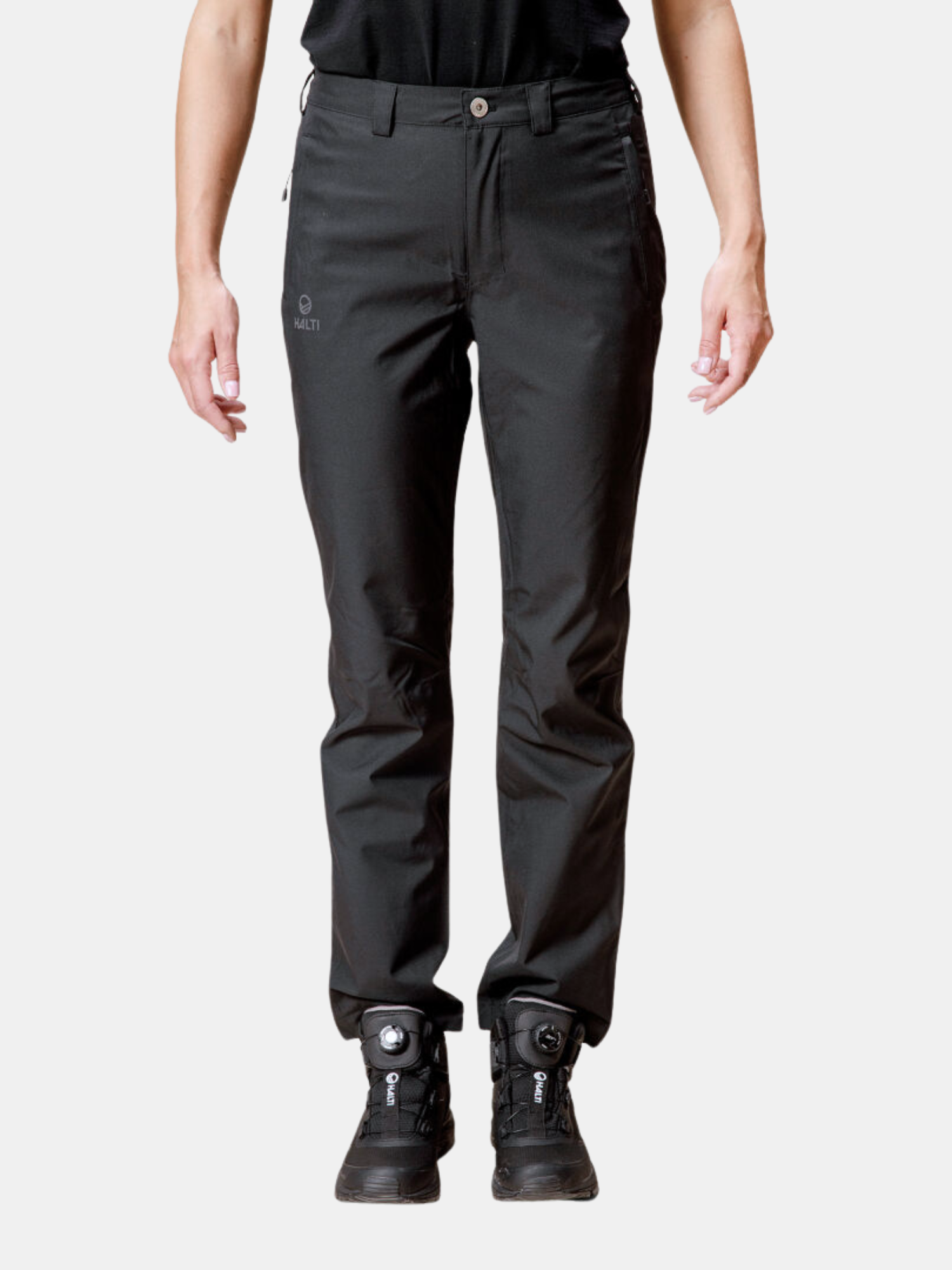 Henkilö, jolla on yllään Halti Leisti DX Kuorihousut Naisten ja mustat saappaat, seisoo vaaleaa taustaa vasten.