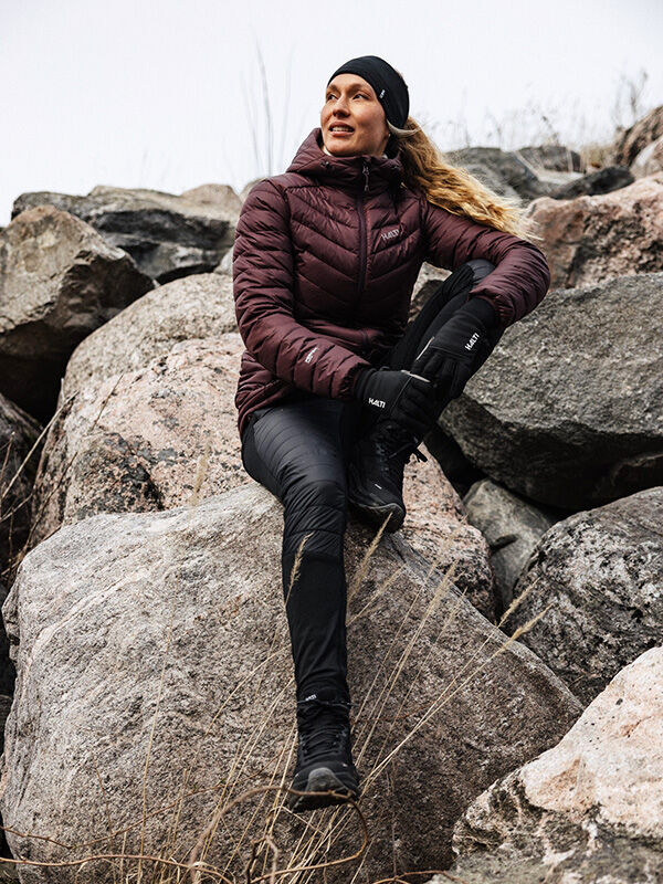 Henkilö ulkoiluvaatteissa ja Halti Veidnes Kevyesti Topatut Housut Naisten istuu suurella kalliolla katsellen sivulle.