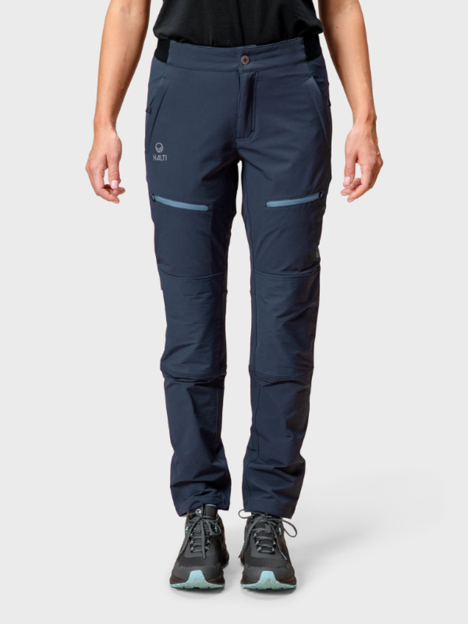 Henkilö, jolla on laivastonsininen Halti Pallas II Naisten Warm X-stretch Housut, musta paita ja tiilenpunaiset kengät, seisoo tavallista taustaa vasten.