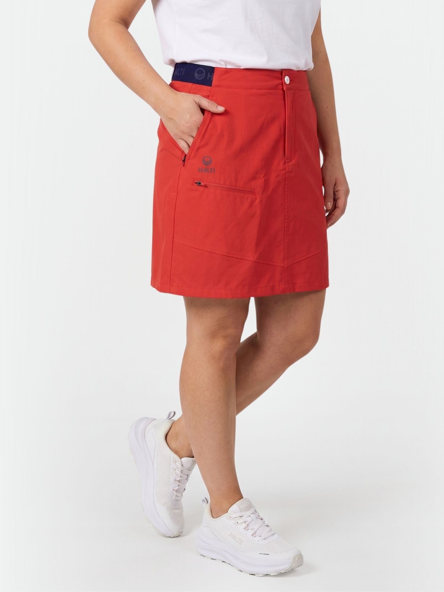 Henkilö, jolla on punainen Halti Pallas Naisten Lite X-stretch Shortsihame, valkoinen t-paita ja lenkkarit, seisoo käsi taskussa.