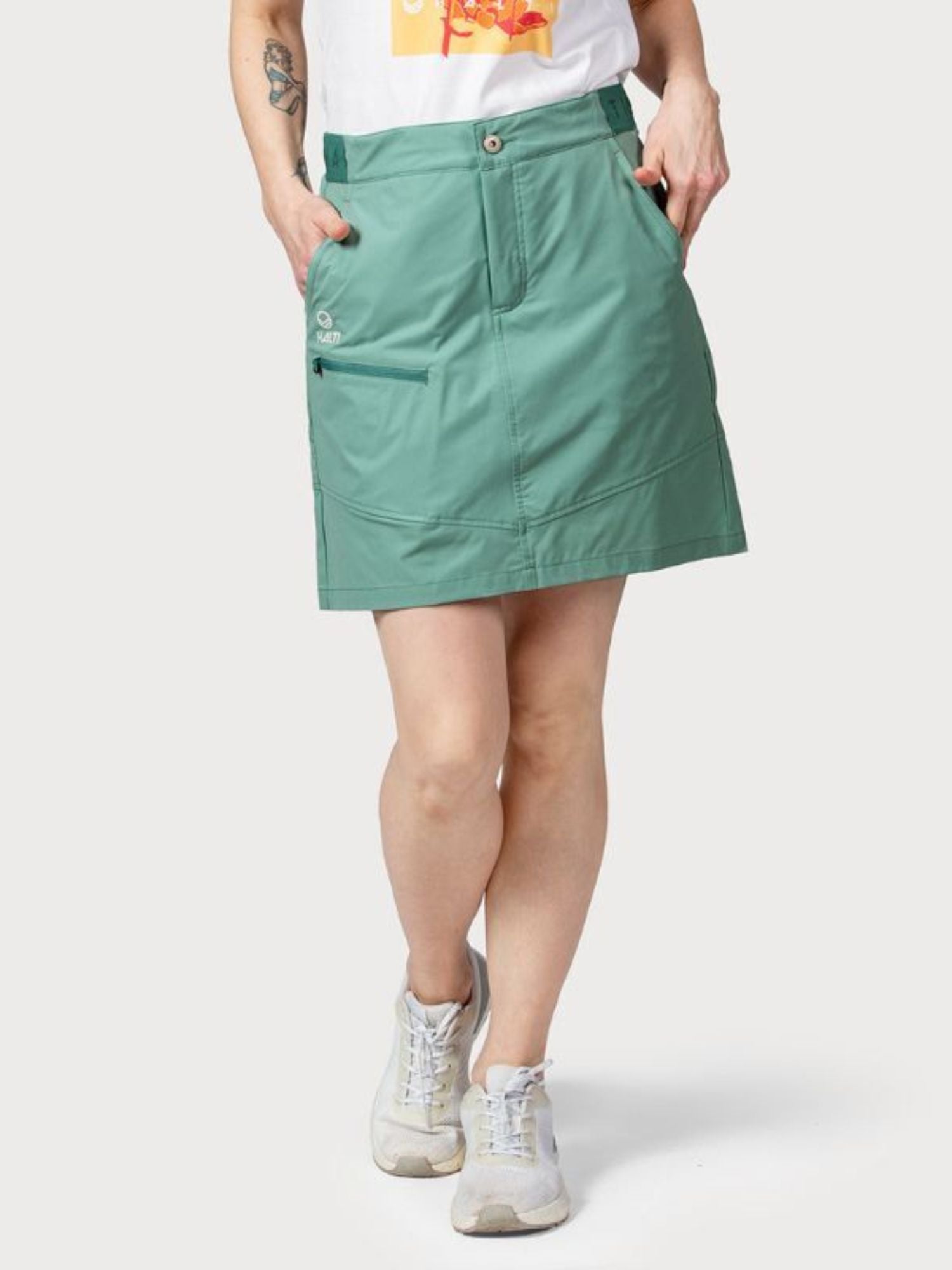 Henkilöllä on vihreät Halti Pallas Naisten Lite X-stretch -shortsit, valkoinen printti-t-paita ja valkoiset lenkkarit, kädet taskuissa.