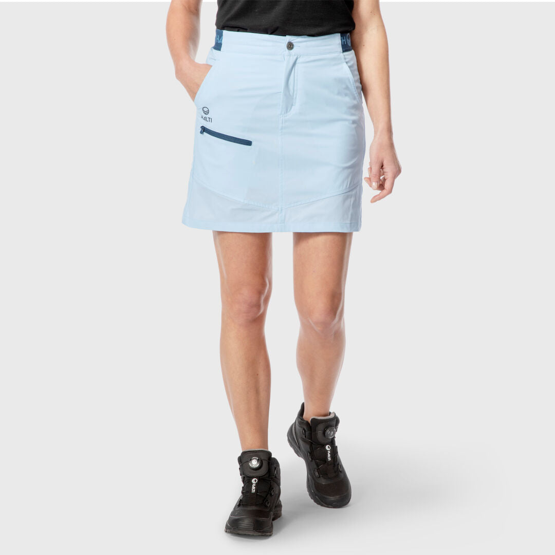 Henkilö vaaleansinisessä Halti Pallas Naisten Lite X-stretch Shortsihameessa, mustassa paidassa ja vaelluskengissä seisoo harmaalla.