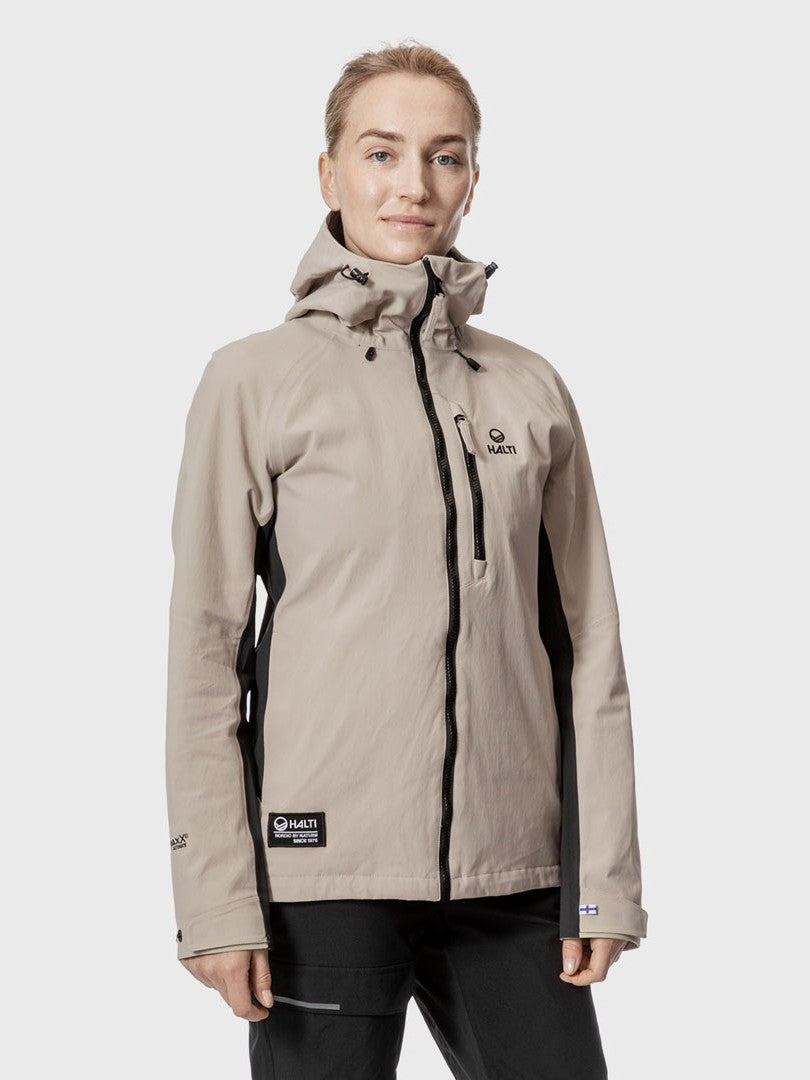 Henkilö käyttää Halti Hiker Naisten DrymaxXX Pro ulkoilutakkia hupulla, beige ja musta, tavallista taustaa vasten.