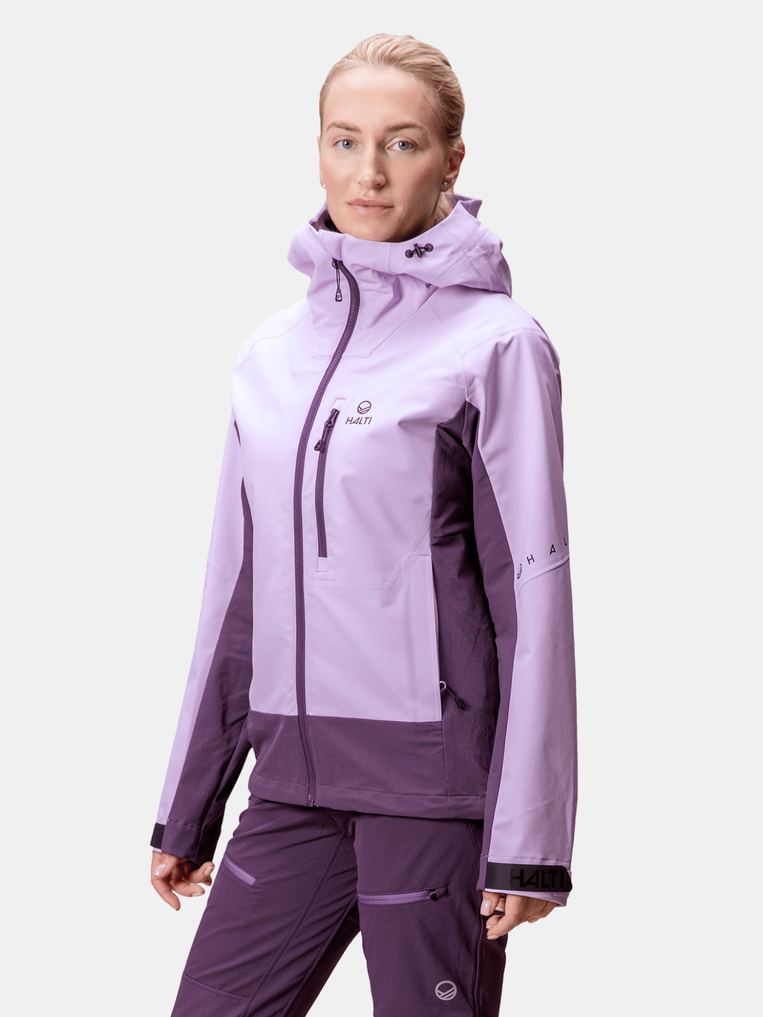 Henkilö seisoo valkoista taustaa vasten pukeutuneena kaksisävyiseen violettiin Halti Pallas III Naisten Warm Hybriditakki -takkiin.